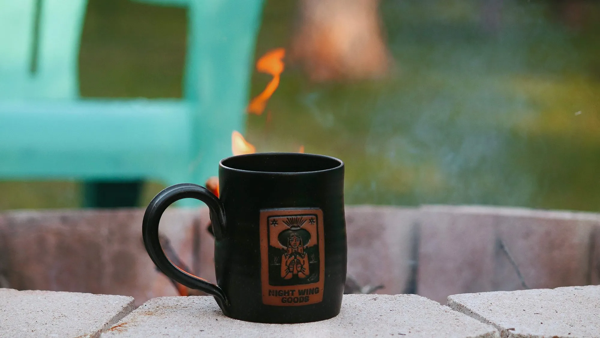 A Night Wing Goods mug beside a fire pit